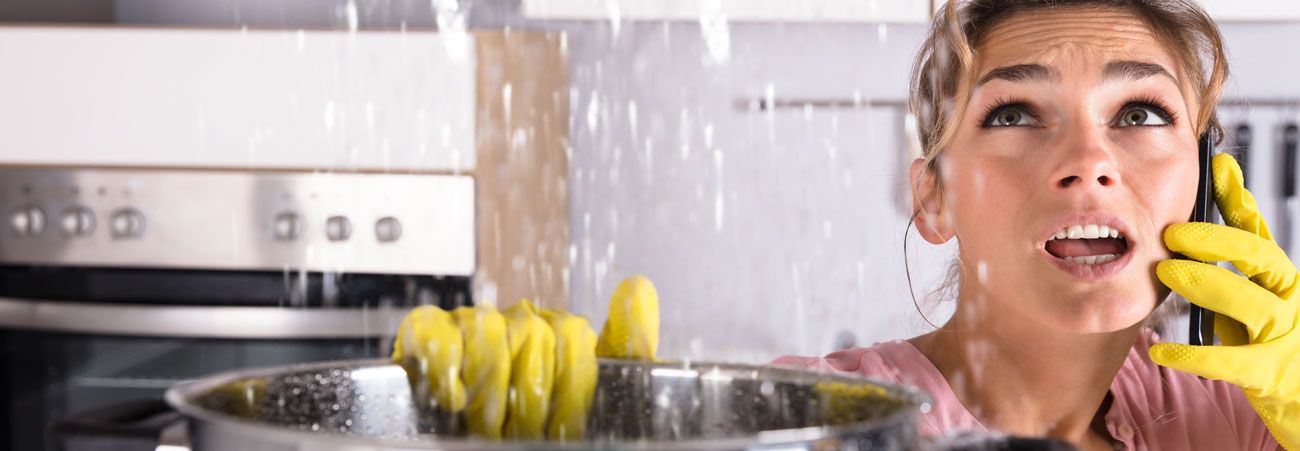 Frau in Küche telefoniert, während Wasser durch die Decke tropft und sie einen Topf zum Auffangen hält.